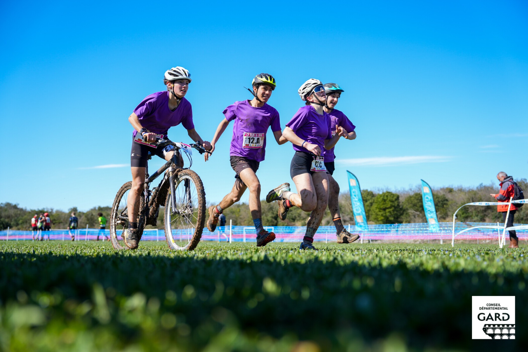 Centre Sportif du Gard : run and bike Photo de 3 jeunes athlètes, 1 en VTT, les 2 autres en course à pied sur une pelouse bien verte et un ciel bleu azur